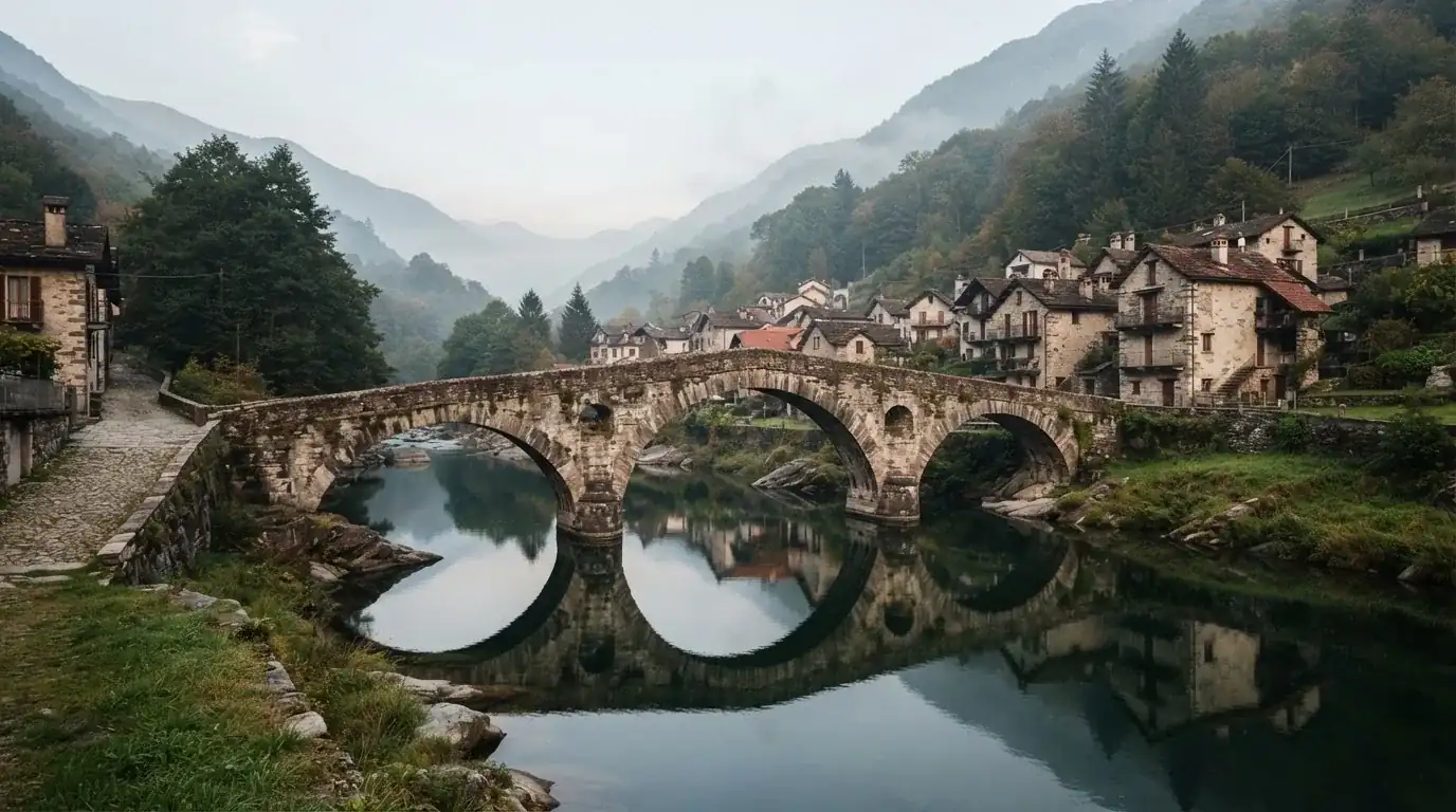 Eine alte Steinbrücke über einen ruhigen Fluss, dahinter Häuser mit Ziegeldächern und bewaldete Hügel.