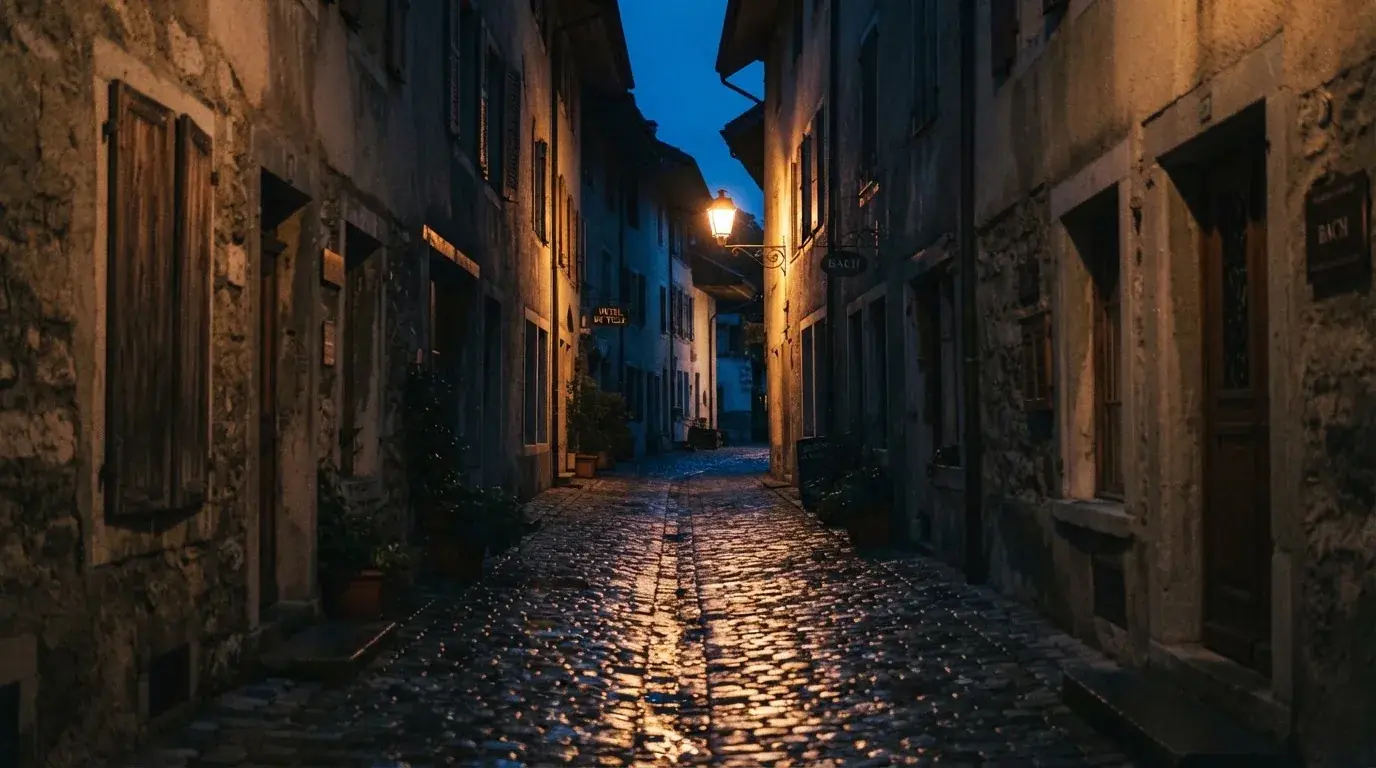 Eine Strassenlaterne in einer leeren Gasse am Abend, das nasse Kopfsteinpflaster spiegelt das warme Licht.