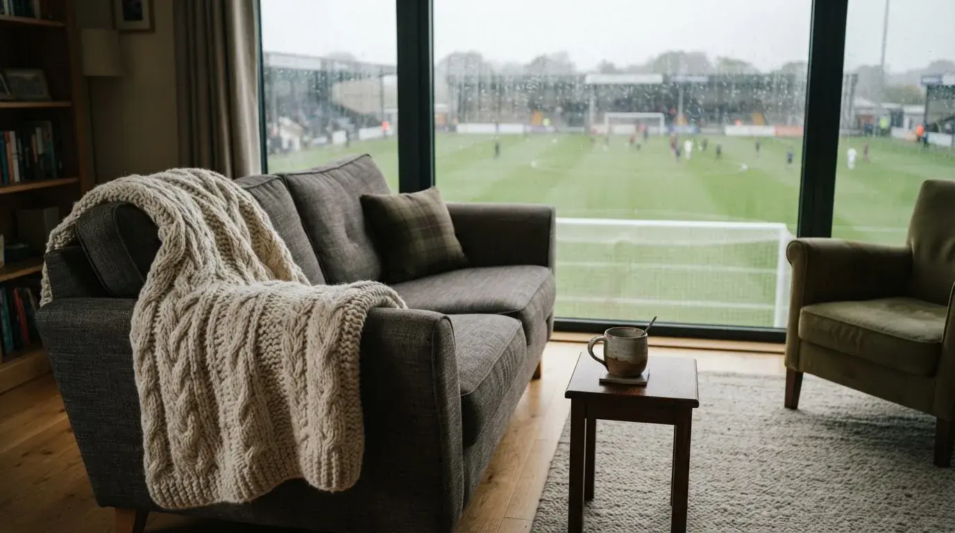 Ein Sofa mit Wolldecke steht vor einem grossen Fenster, draussen Regen und in der Ferne ein Fussballplatz.
