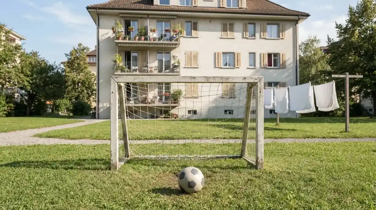 Ein kleines Holztor und ein Ball stehen auf einer Wiese hinter einem Mehrfamilienhaus, die Wäscheleine hängt im Wind.