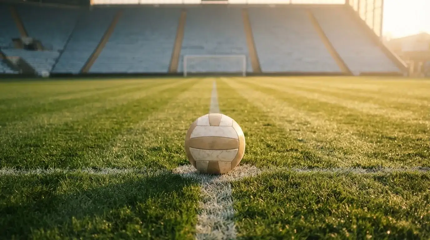 Ein Fussball liegt auf dem Anstosspunkt eines leeren Rasenplatzes im frühen Morgenlicht, dahinter erhebt sich eine menschenleere Tribüne.