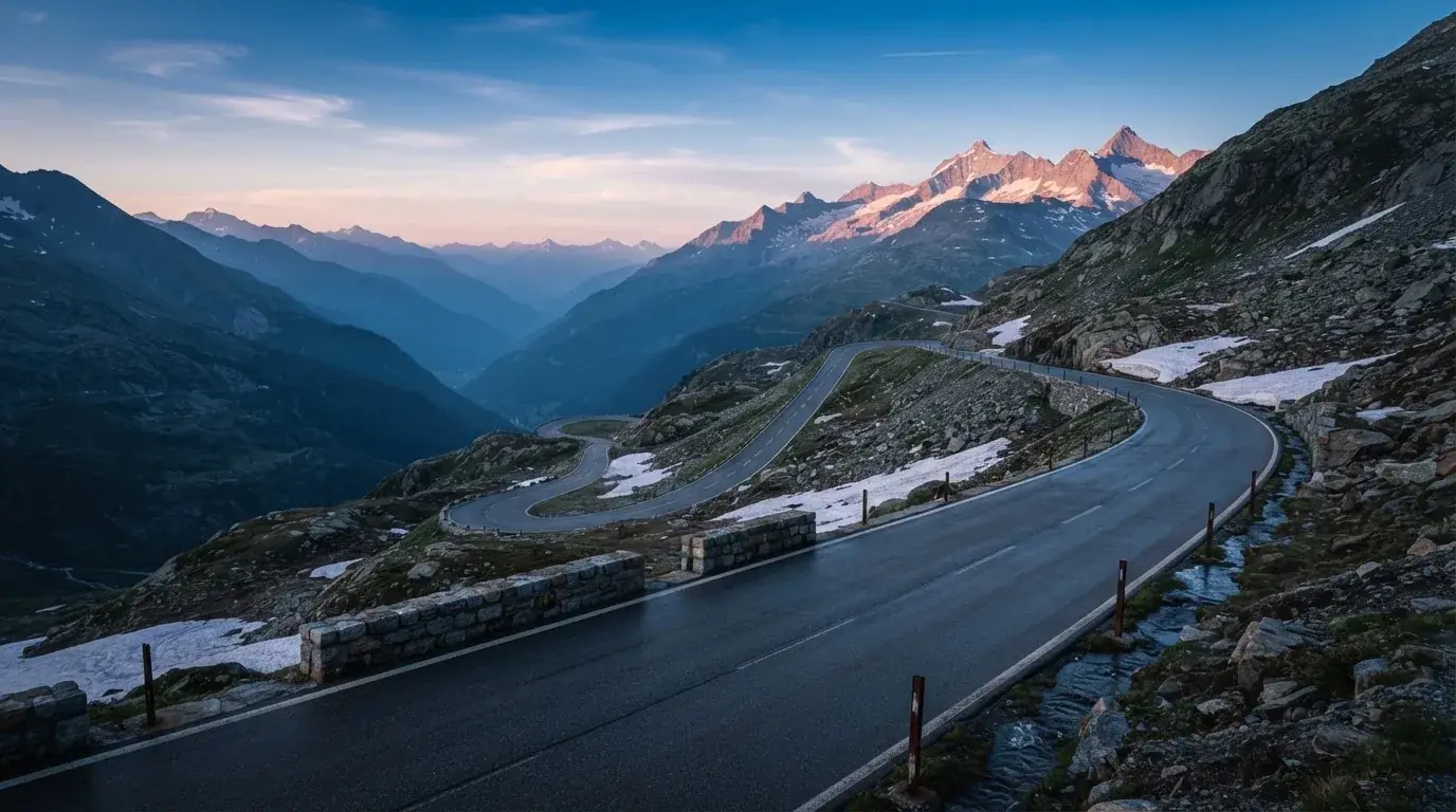Ein Bergpass in den Schweizer Alpen mit leerer Strasse und Schneeresten am Rand im ersten Frühlicht.