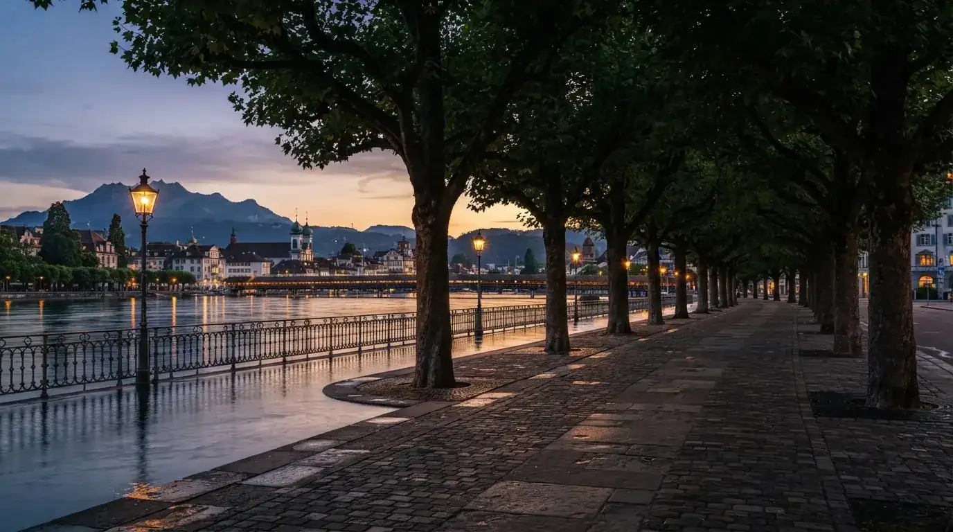 Eine Rheinpromenade mit hohen Bäumen am Abend, das Wasser spiegelt das Licht der ersten Laternen.