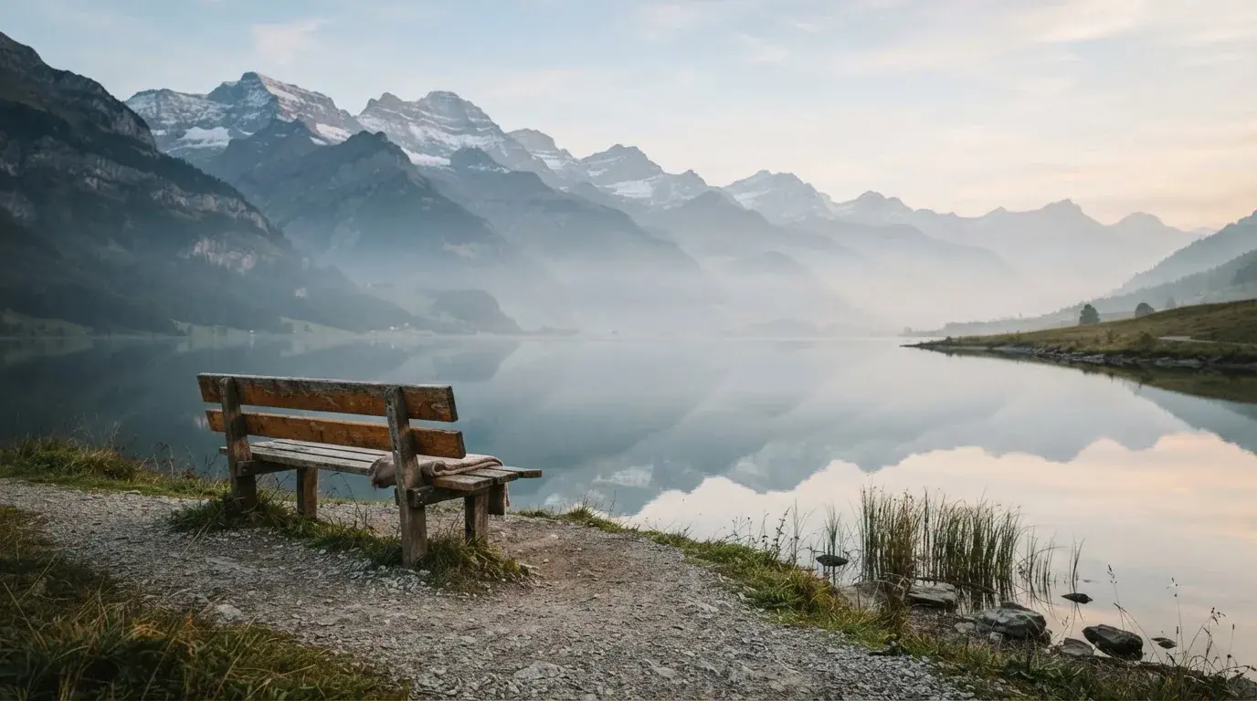 Eine Holzbank an einem Seeufer mit Blick auf eine Bergkette im Morgendunst, das Wasser ist spiegelglatt.