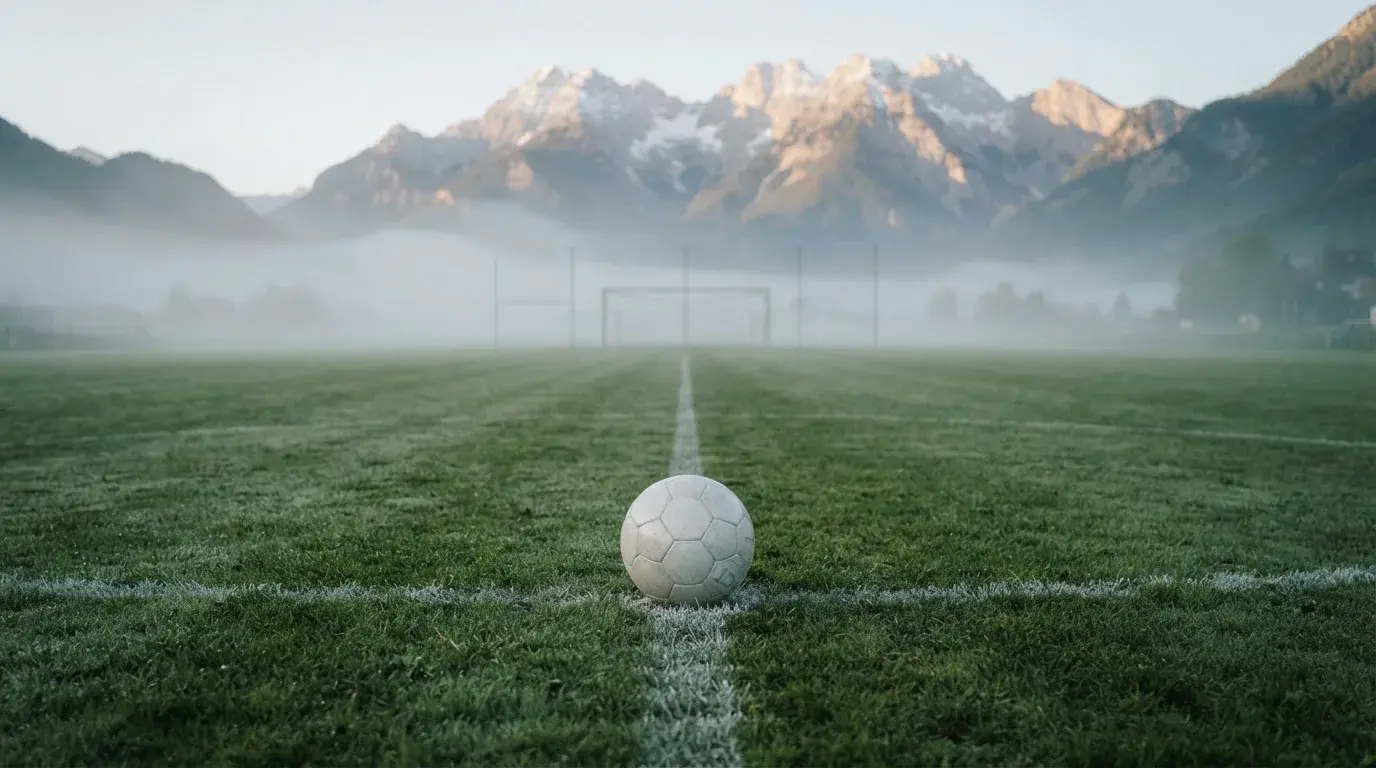 Ein einzelner Fussball auf dem Mittelpunkt eines leeren Rasens im Morgennebel, hinter dem Tor ragen Berge auf