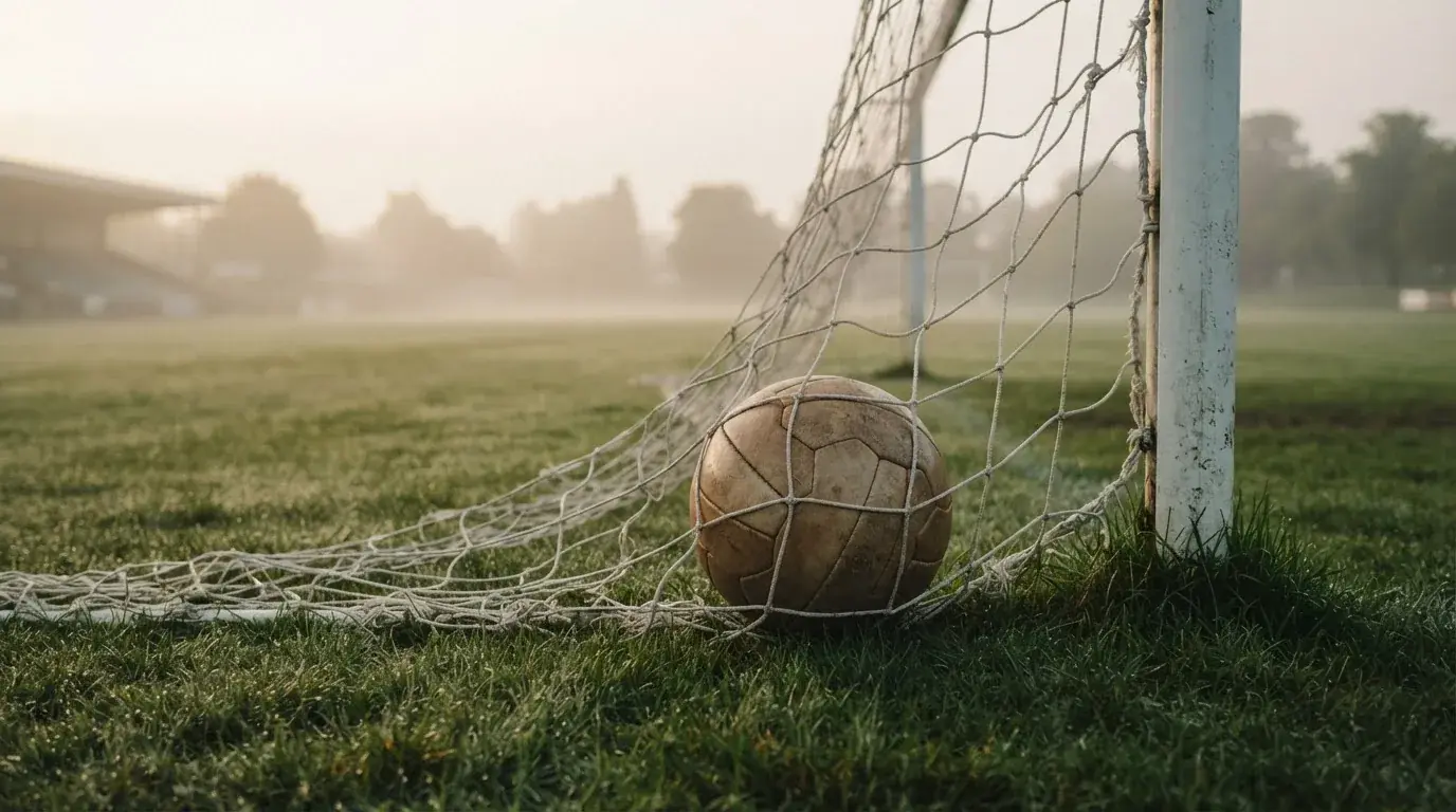Ein Fussball liegt im Netz hinter einem Tor, weicher Morgennebel hängt über dem Rasen