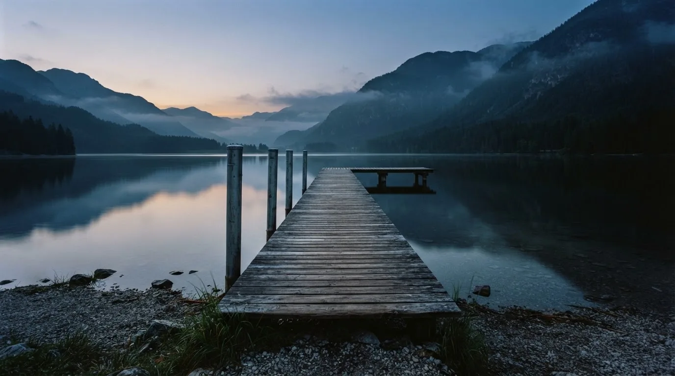 Ein Bergsee in der Dämmerung mit einem leeren Holzsteg, die Wasseroberfläche ist ruhig und Nebel zieht über die Hänge.