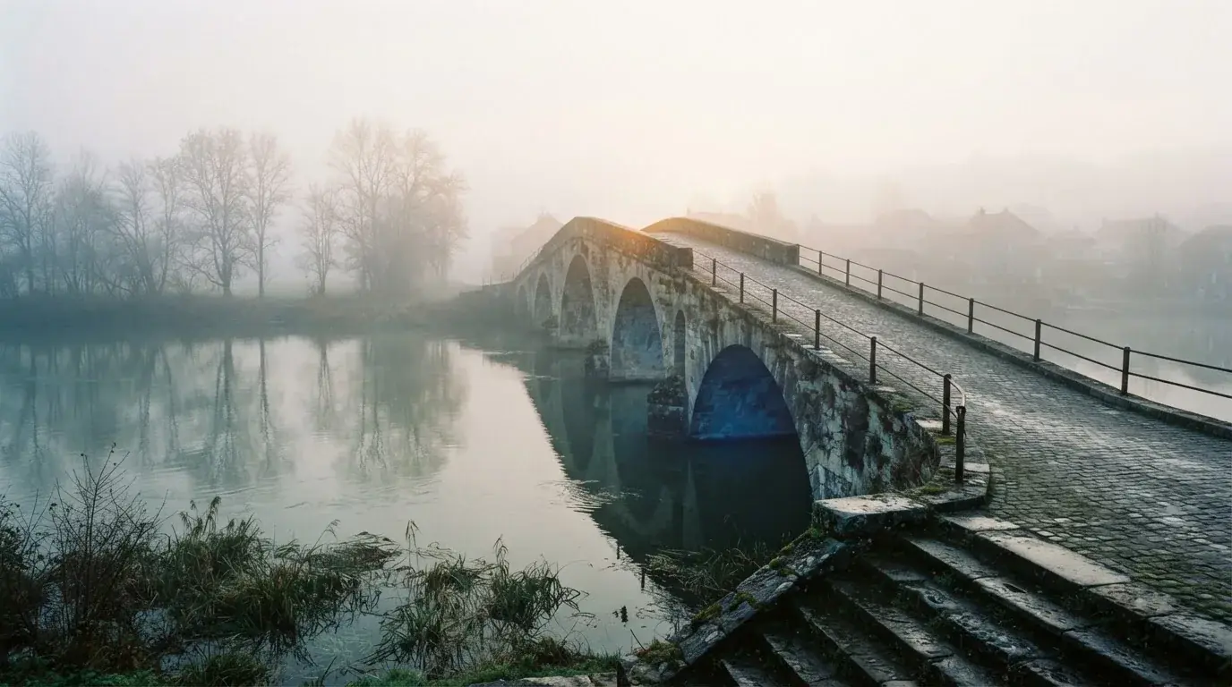 Eine Steinbrücke führt im Frühnebel über einen Fluss, auf dem Fussweg ist niemand zu sehen.